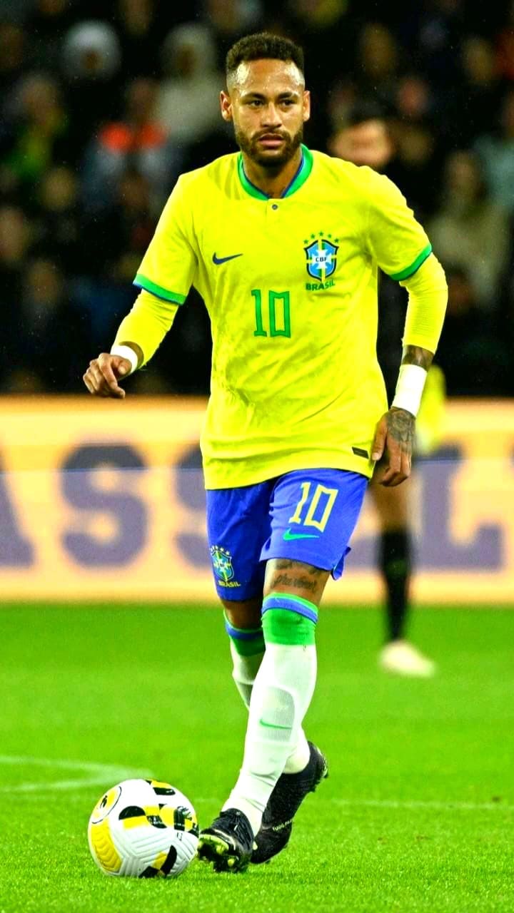 A soccer player in a yellow Brazil jersey with the number 10 and blue shorts is dribbling a ball on the field during a match, while spectators and players fill the blurred background—perfect action to catch live on IPTV Latino.